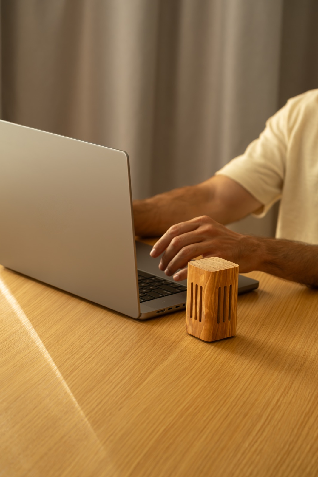 A square olive wood diffuser on a desk beside someone working on a laptop